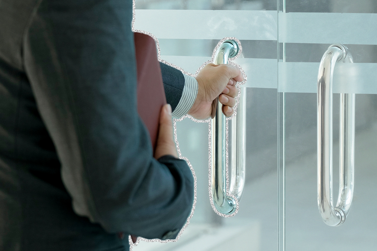A person in a business suit with a briefcase in one hand and opening a glass door with the other hand.