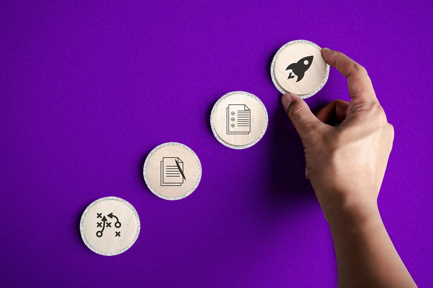 A purple background with four wooden coins positioned to ascend from the bottom left to top right. A person's hand holds the top coin that has the image of a rocket on it.